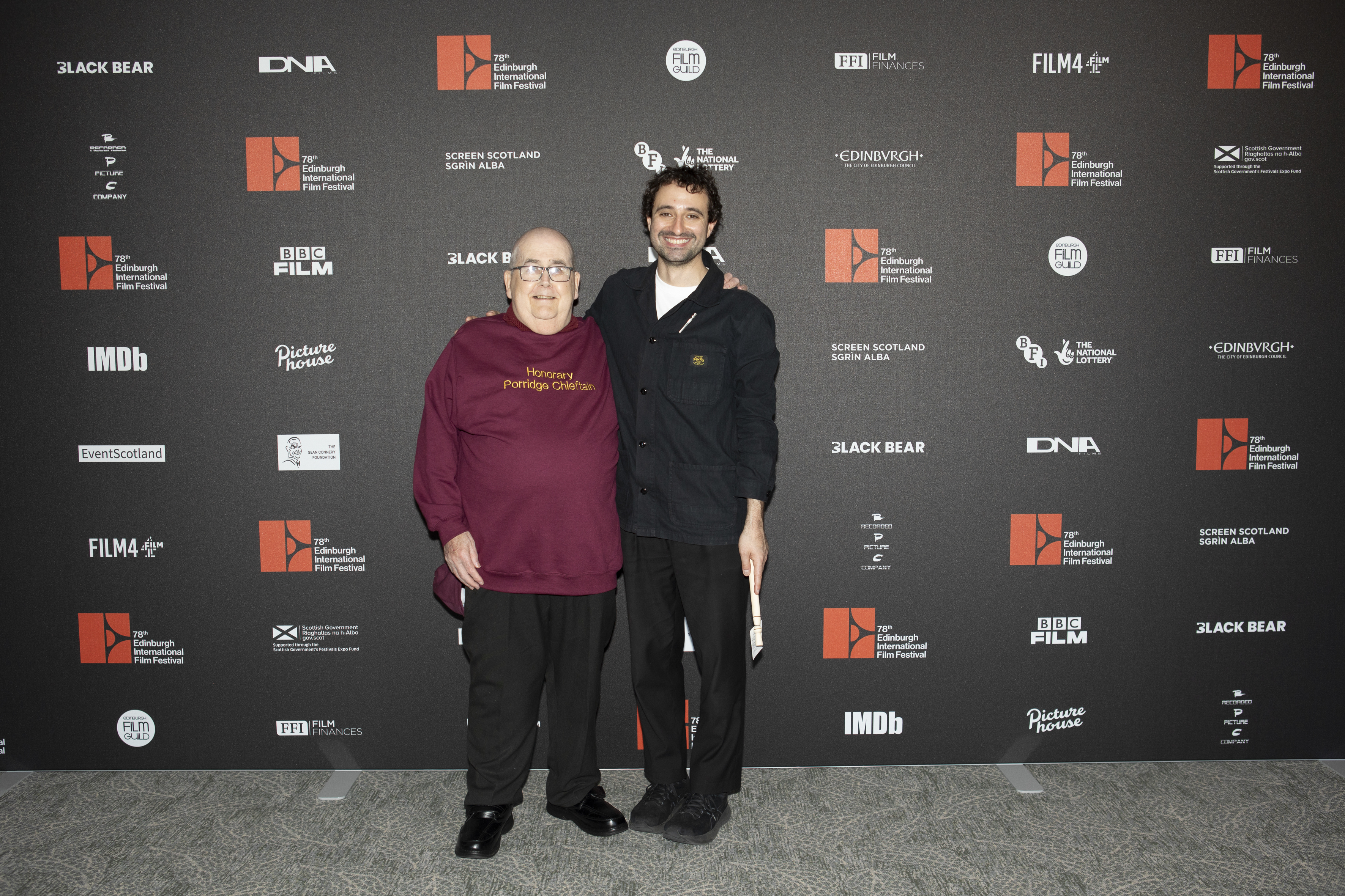 Two men standing in front of a banner of logos at the Edinburgh International Film Festival.