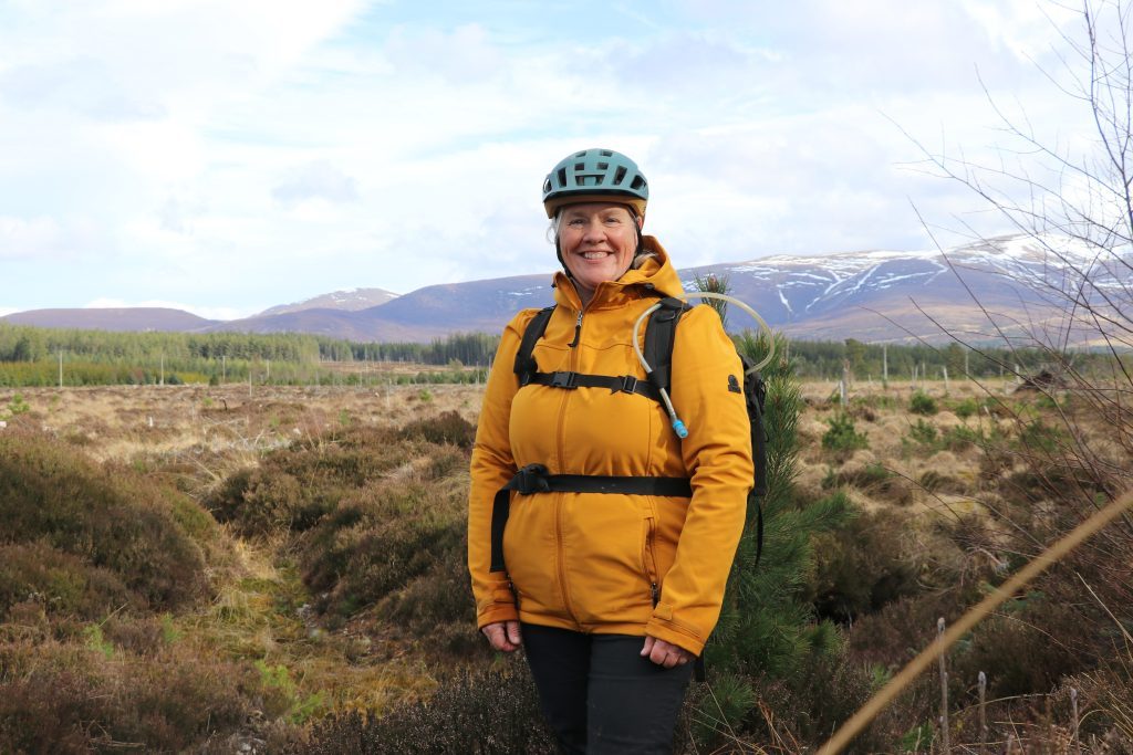 Image of Sarah Rathbone after a Trail Therapy session in the Cairngorms National Park, wearing a yellow jacket, rucksack and cycle helmet and standing amongst heather with a snowy mountain backdrop.