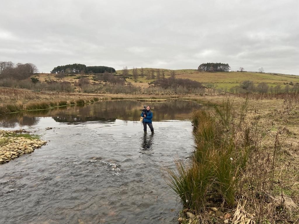 Image of Keilidh Ewan stood in the Beltie Burn on the River Dee holding a camera and microphone on her shoulder.