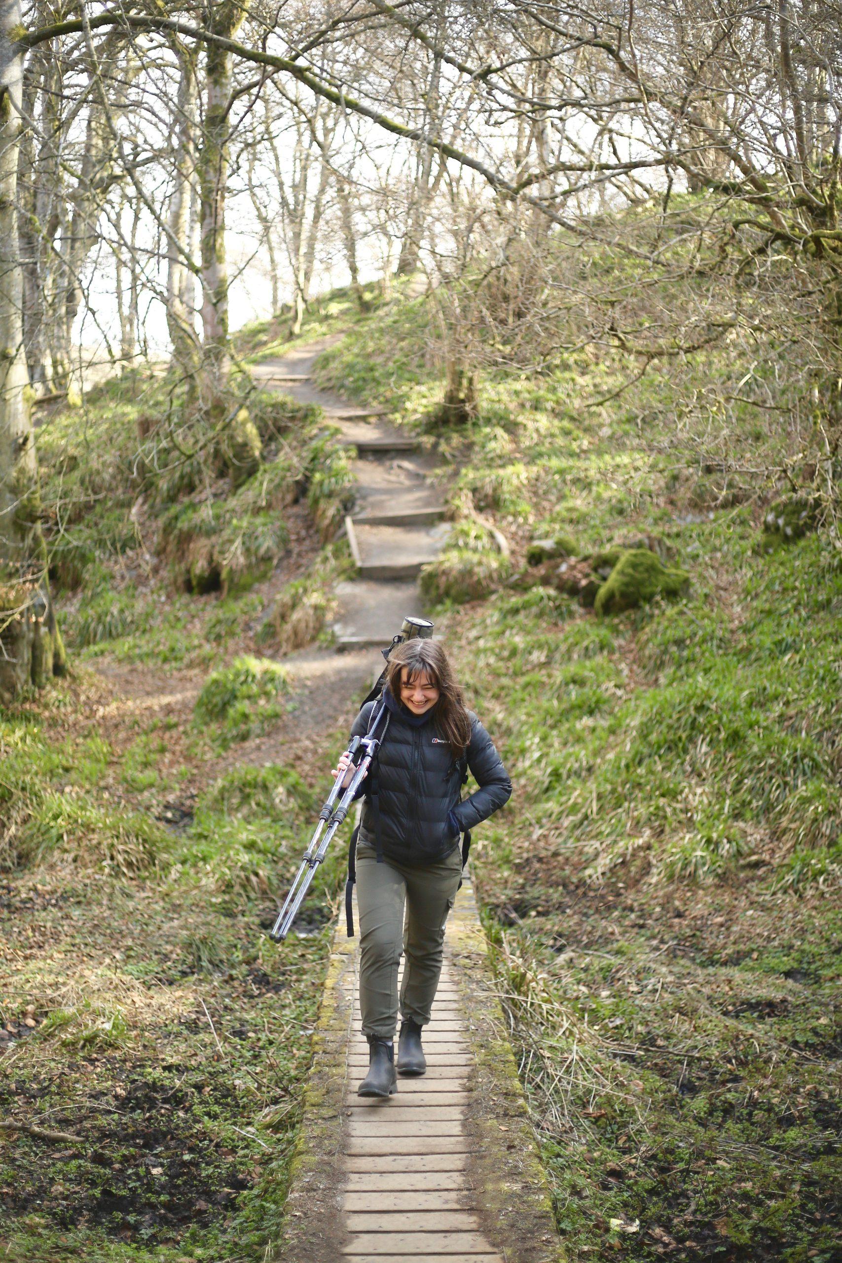 Image of Keilidh Ewan carrying a spotting scope on a tripod across a wooden walkway in woodland.