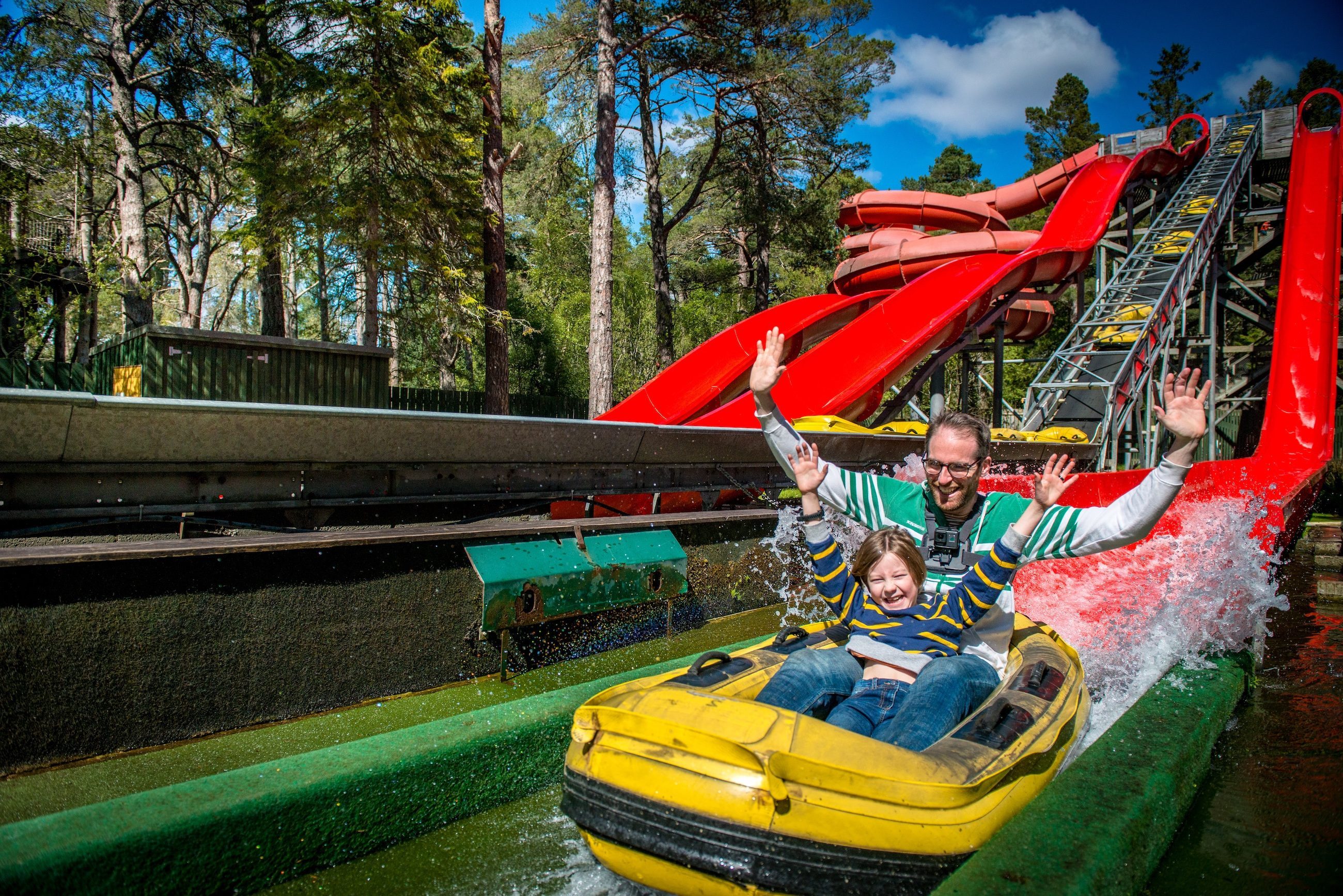 An adult sitting behind a child on a yellow and red water coaster ride with trees in background