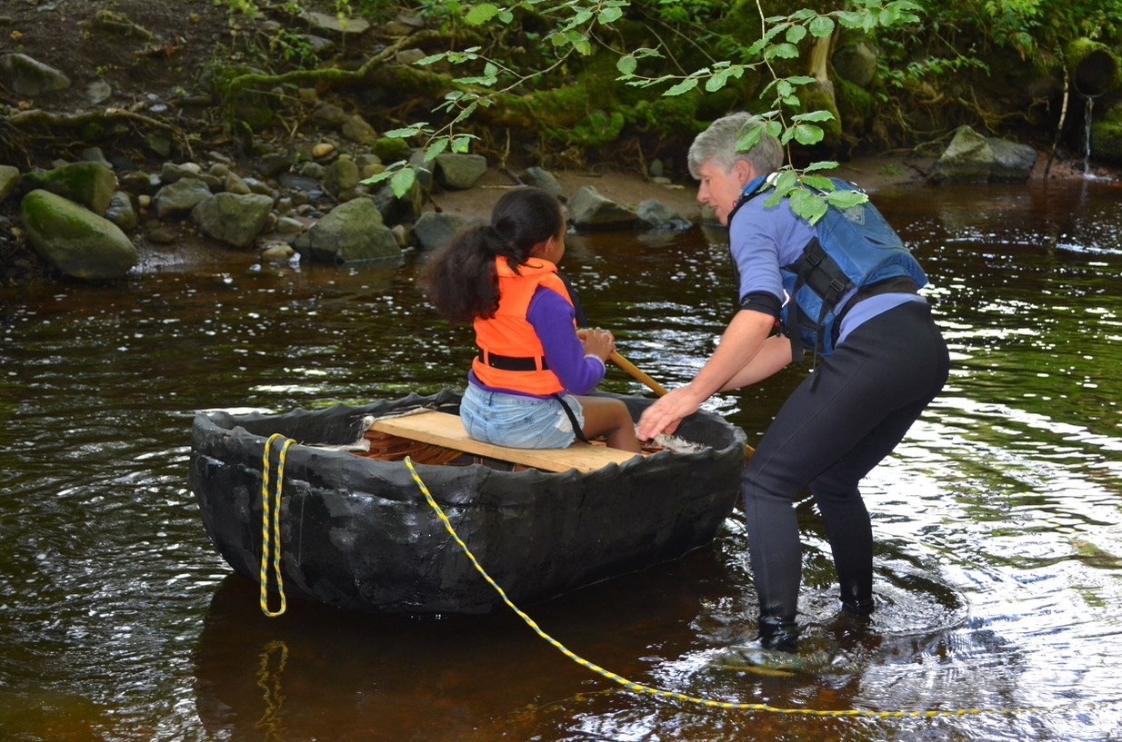 A child being helped to paddle a coracle boat in shallow water.
