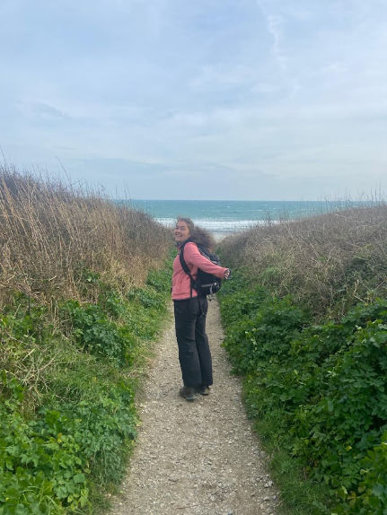 A person standing on a path in a pink jumper with the ocean behind them