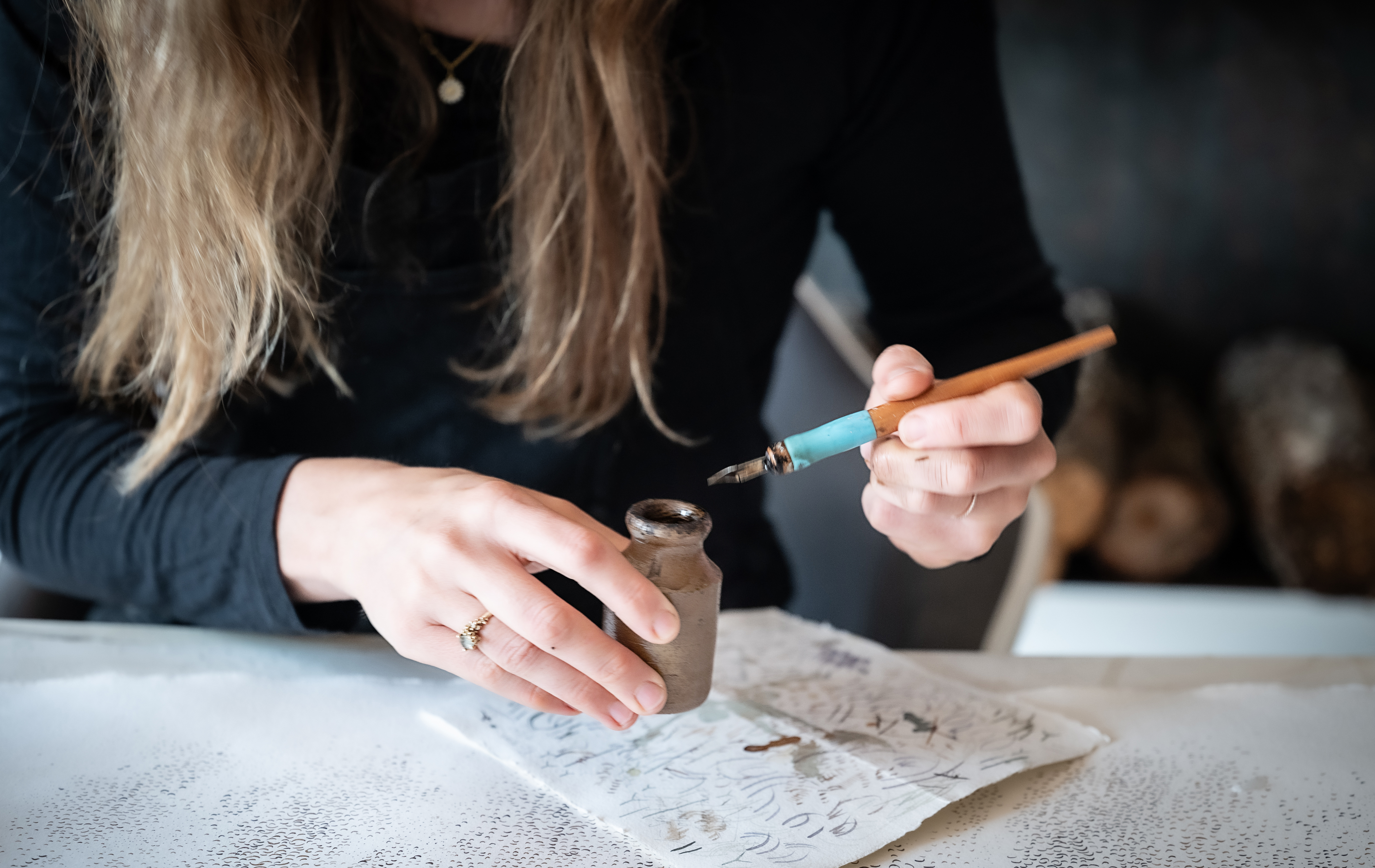 A person holding an ink pen dipping into a vase with paper with scirbbles on it below.