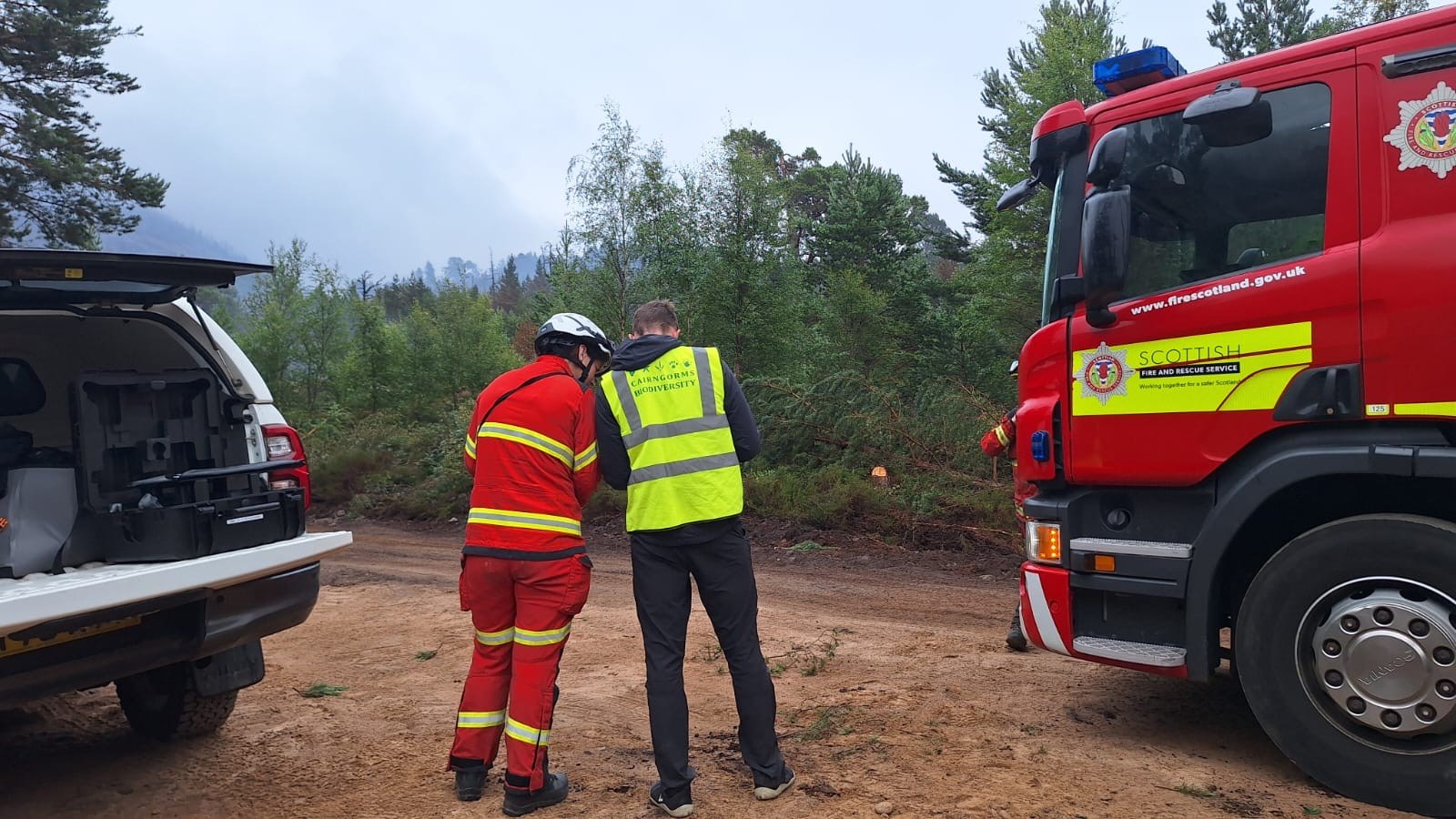two people stand beside fire engine and pickup truck, with smoke in background.