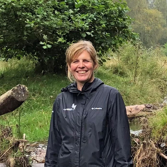 Image of Sally Mackenzie wearing a black jacket and smiling at the camera, stood beside Allt Lorgy burn near Carrbridge.