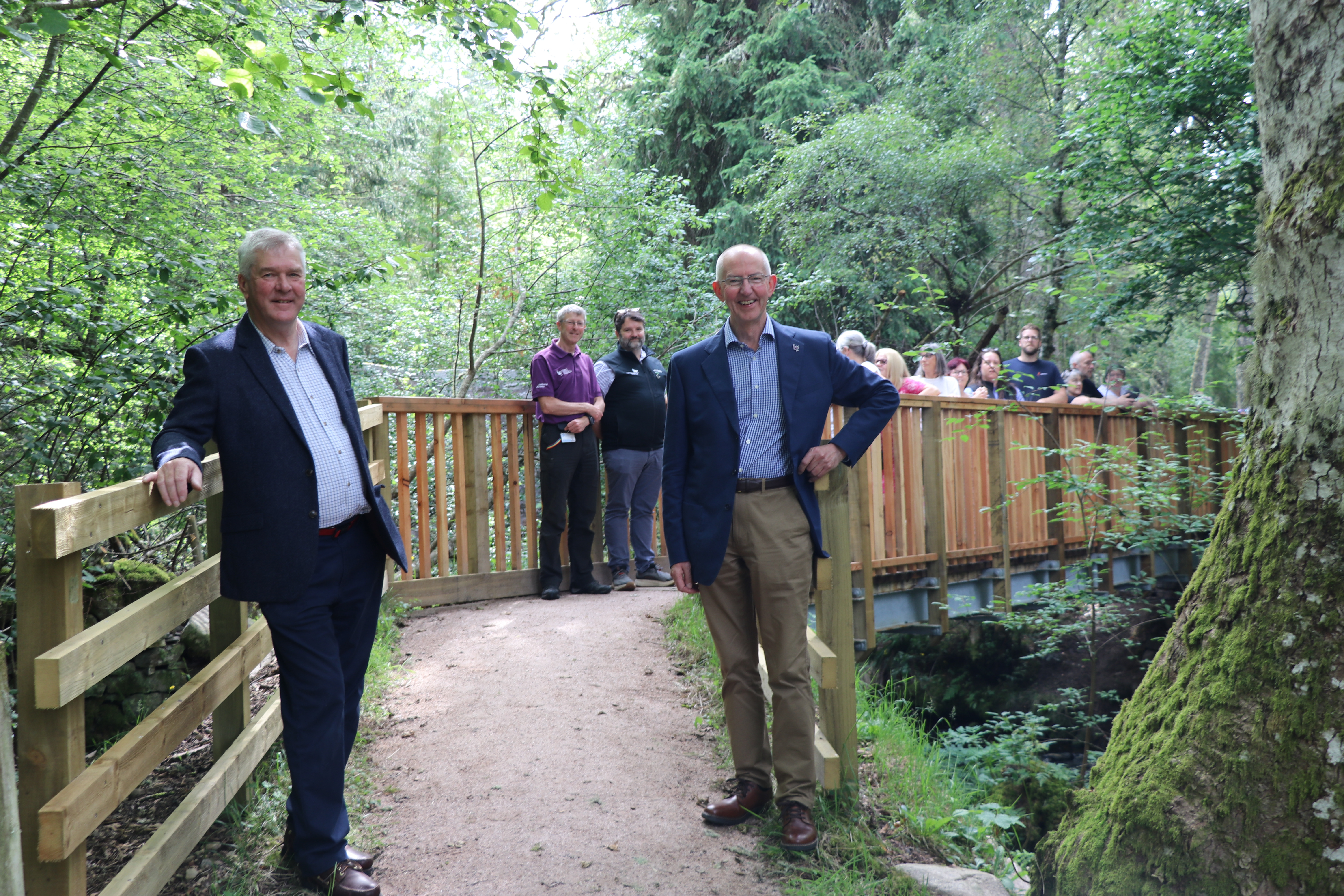 Two men stand in foreground of a bridge, with a group of people in the background.