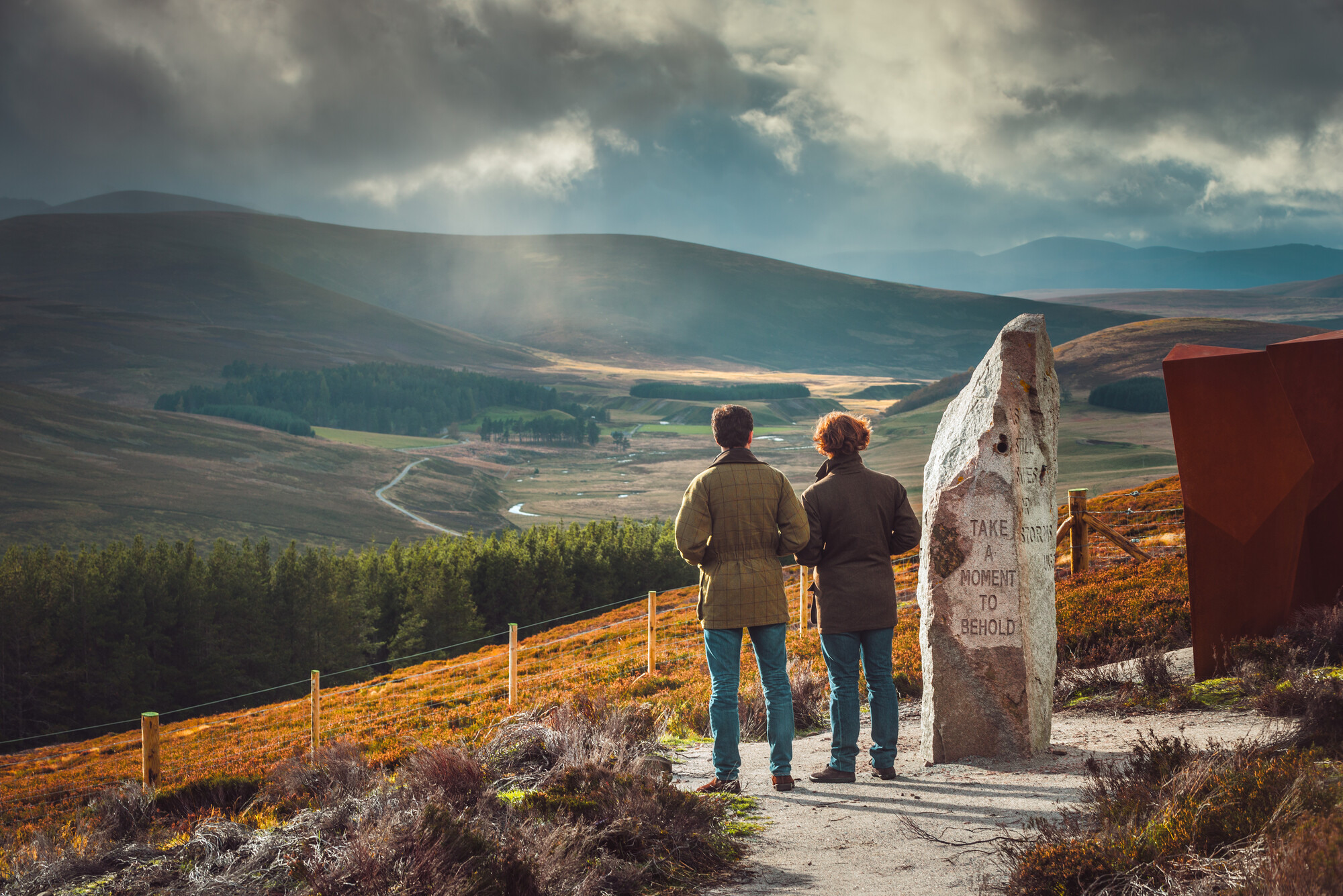 Two people standing at the Watcher viewpoint at Corgarff looking at the view of the glen with trees and hills.