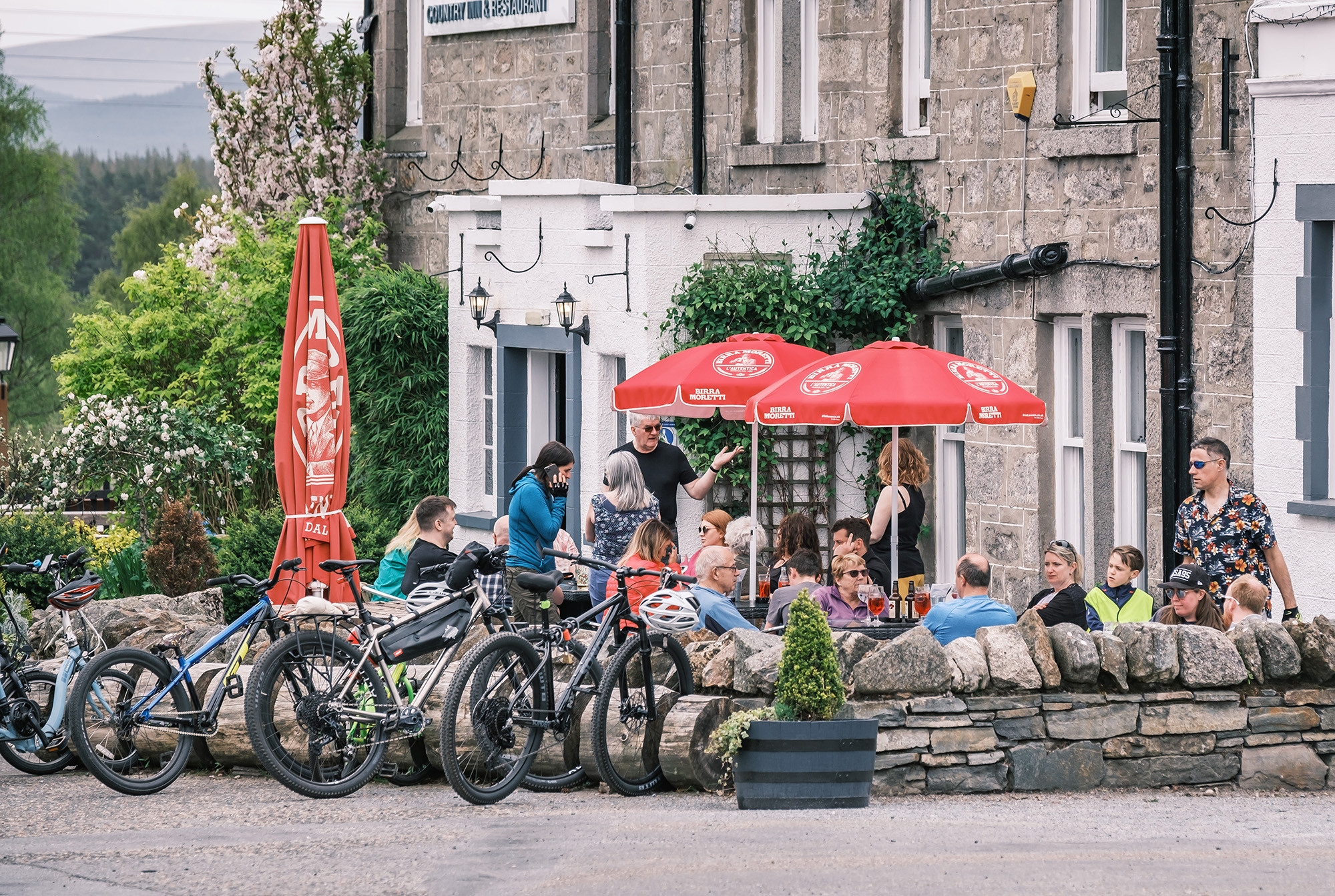 People sitting and standing in a hotel beer garden with red parasols above their heads and bikes in the foreground.