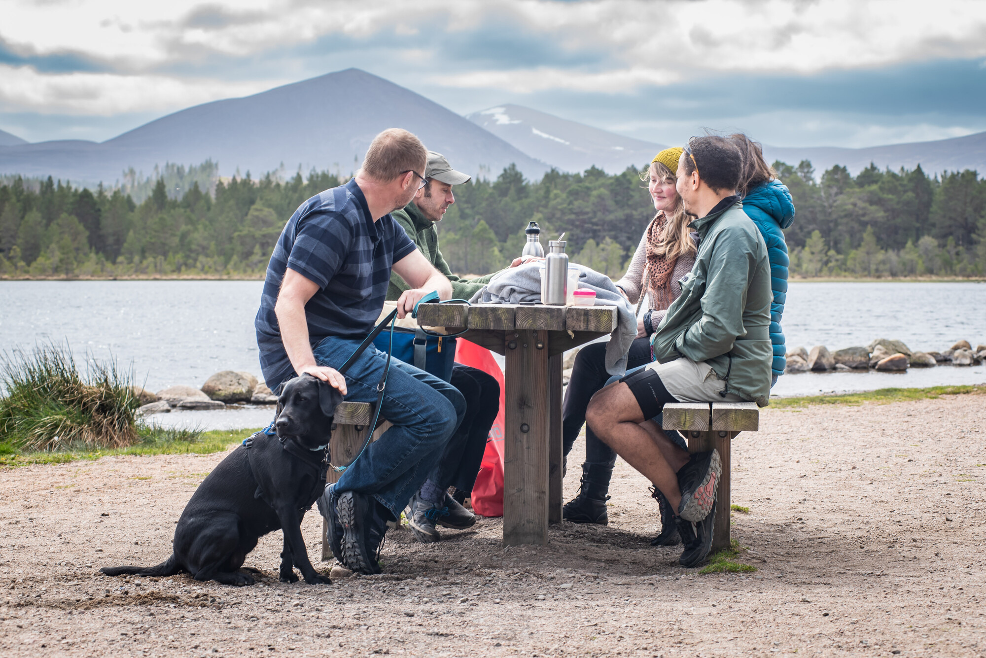 Five people and a dog around a picnic table on the beach at Loch Morlich.