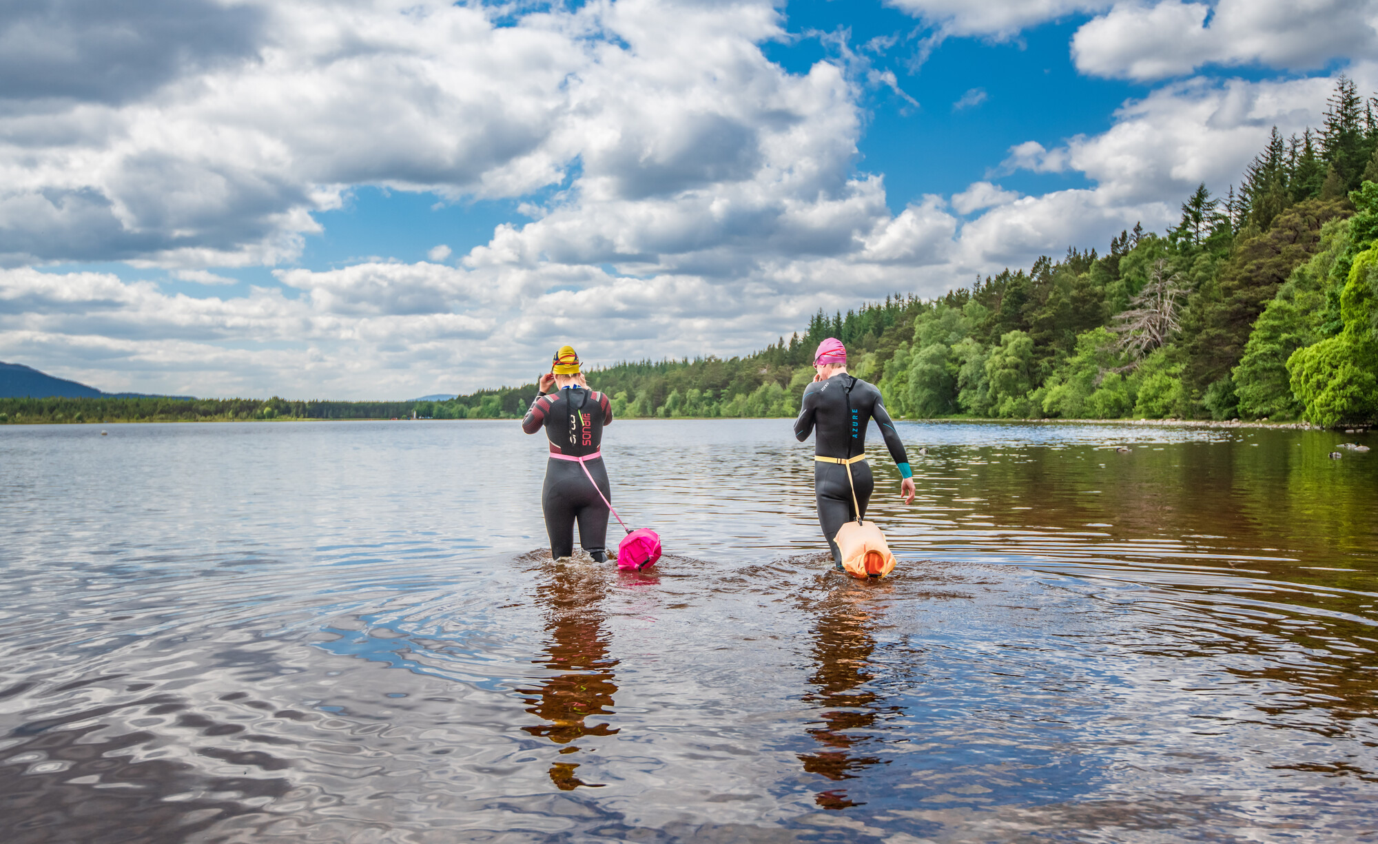 Two people wearing wetsuits with safety floats entering the loch to go swimming.