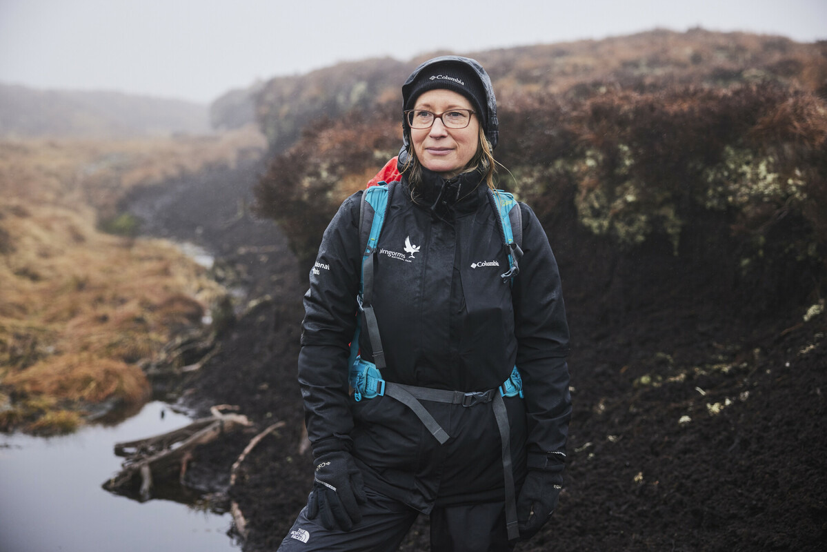 Emma Stewart, wearing a black jacket, blue rucksack and a woolly hat, stood beside some degraded peatland on a misty day