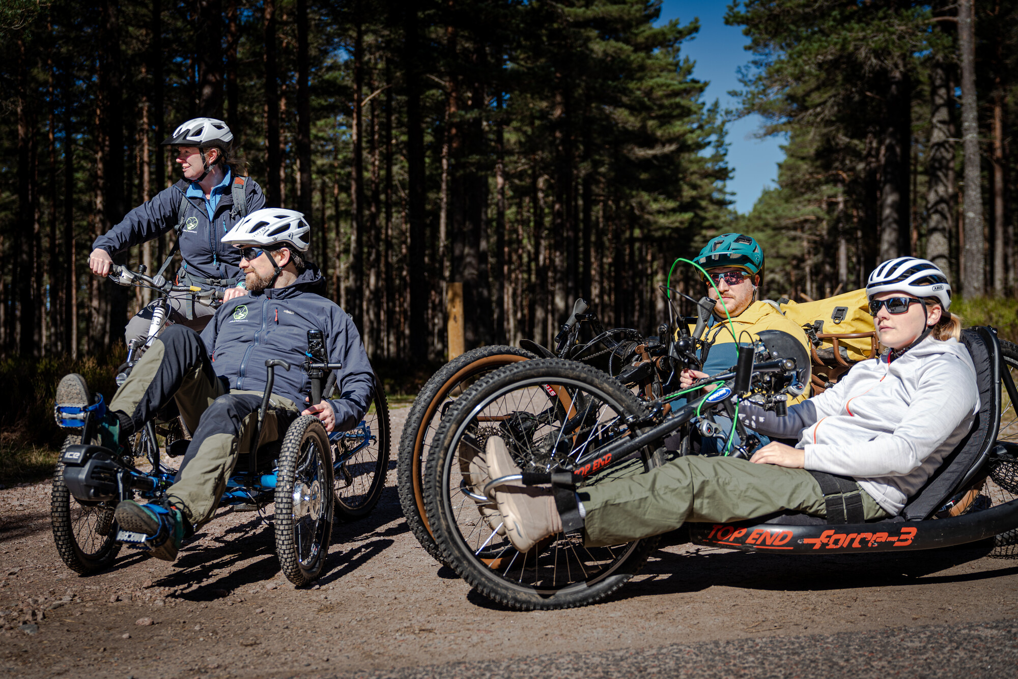 Four people using adaptive bikes and all terrain wheelchairs on forest track.
