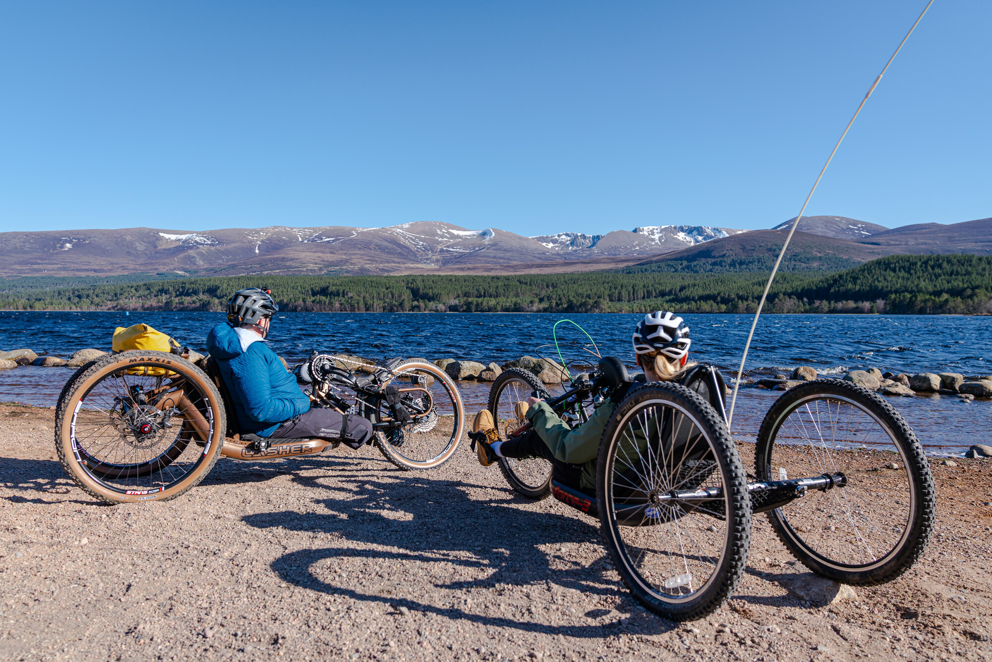 People using adaptive bikes at Loch Morlich beach with loch and mountains in the background.