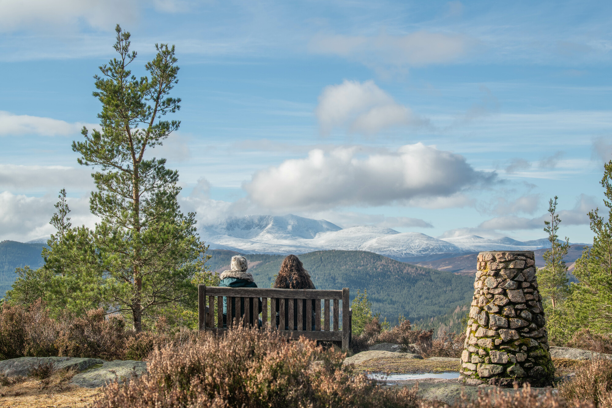 A rock cairn to the right of two women sitting on a bench looking out at a view of trees and hills.