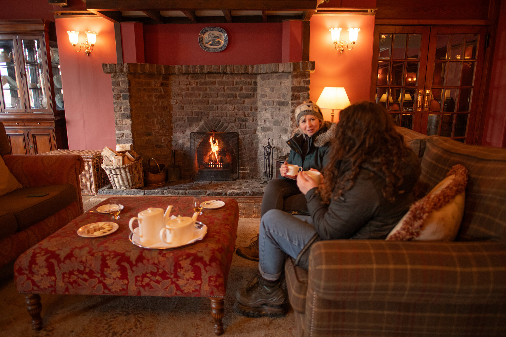Two women sitting on a couch in front of a fireplace with tea and biscuits.