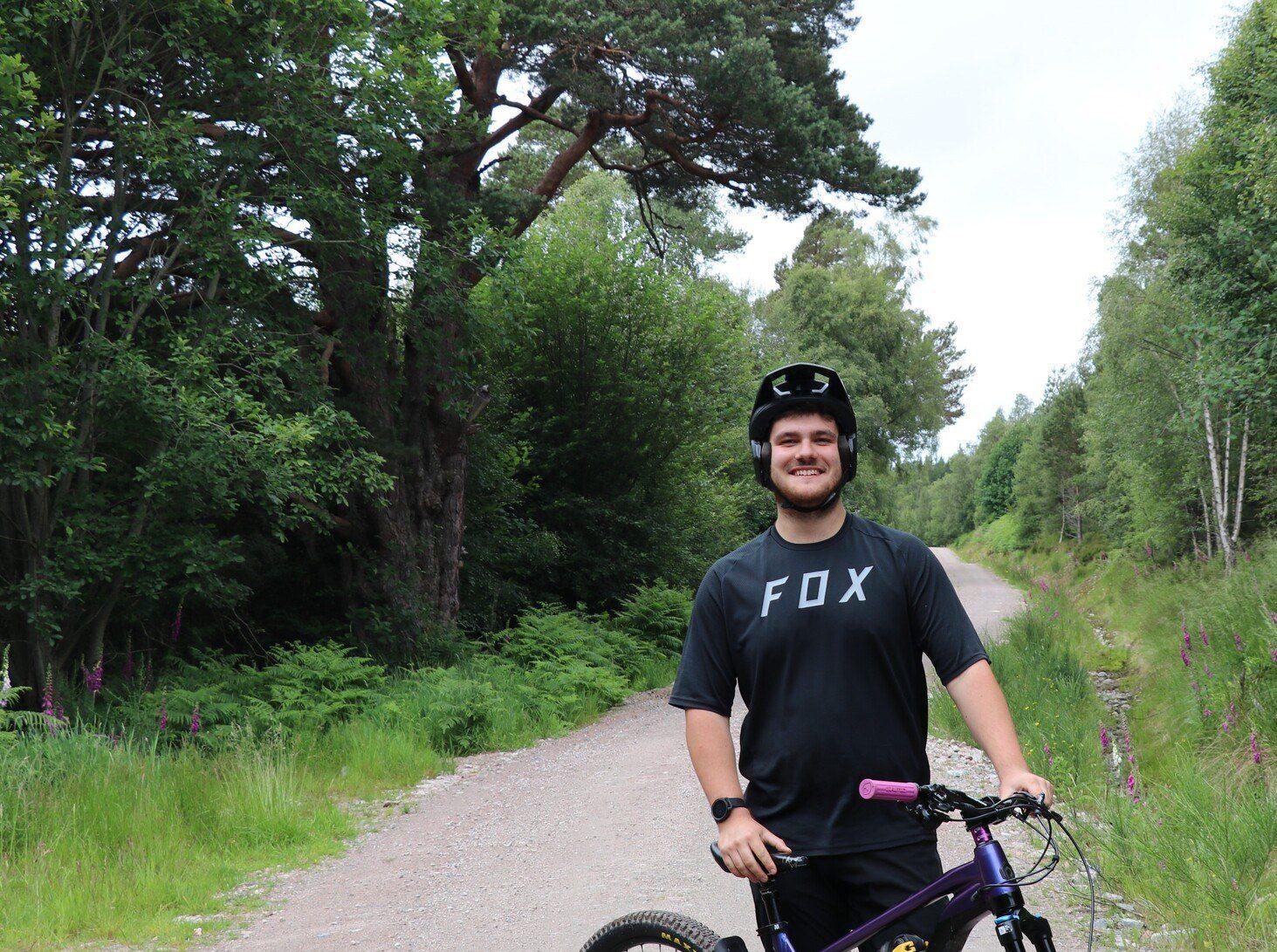 Josh standing with his bike on a wide woodland path.