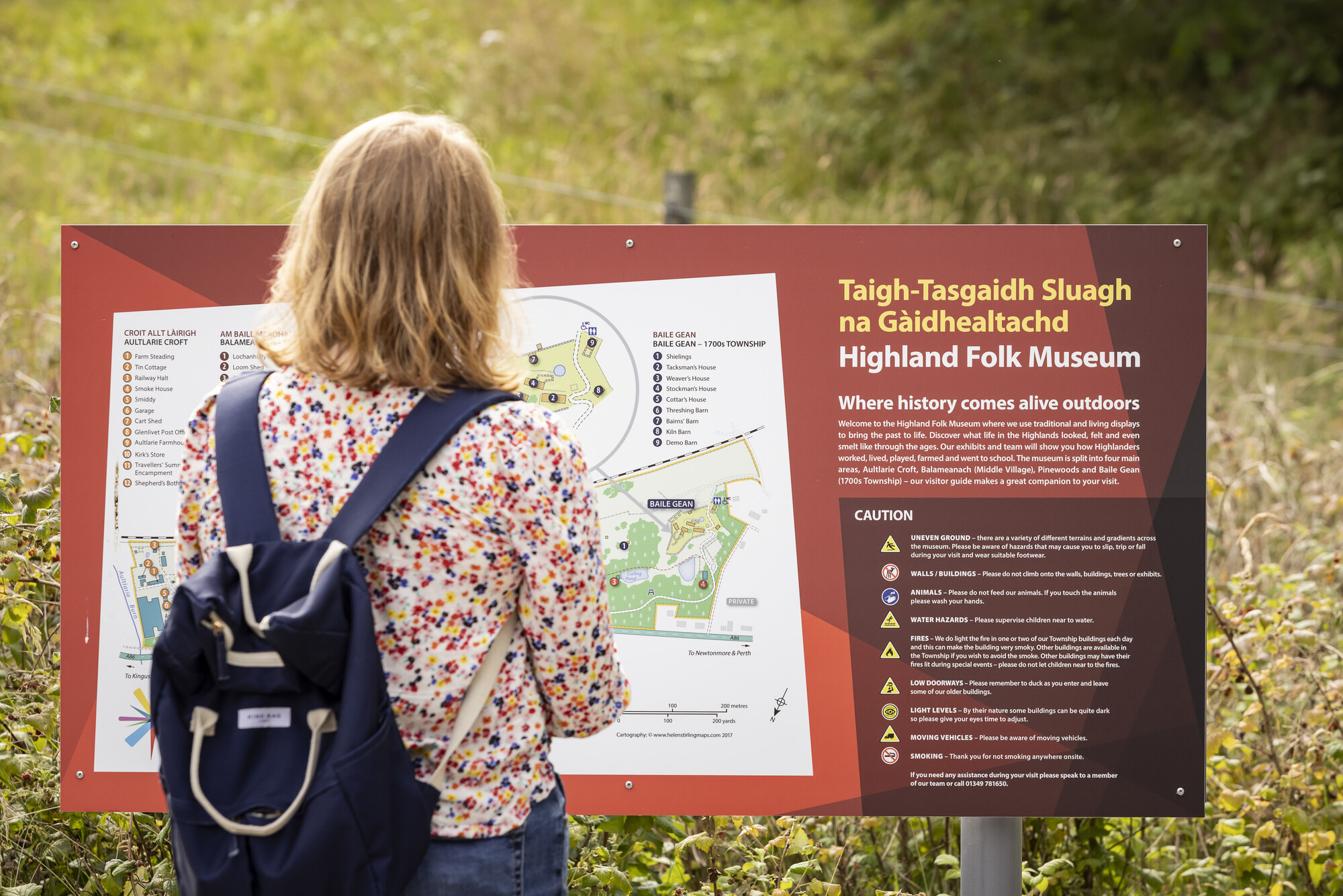 Person standing in front of and reading an interpretation panel with some Gaelic language content at the Highland Folk Museum.