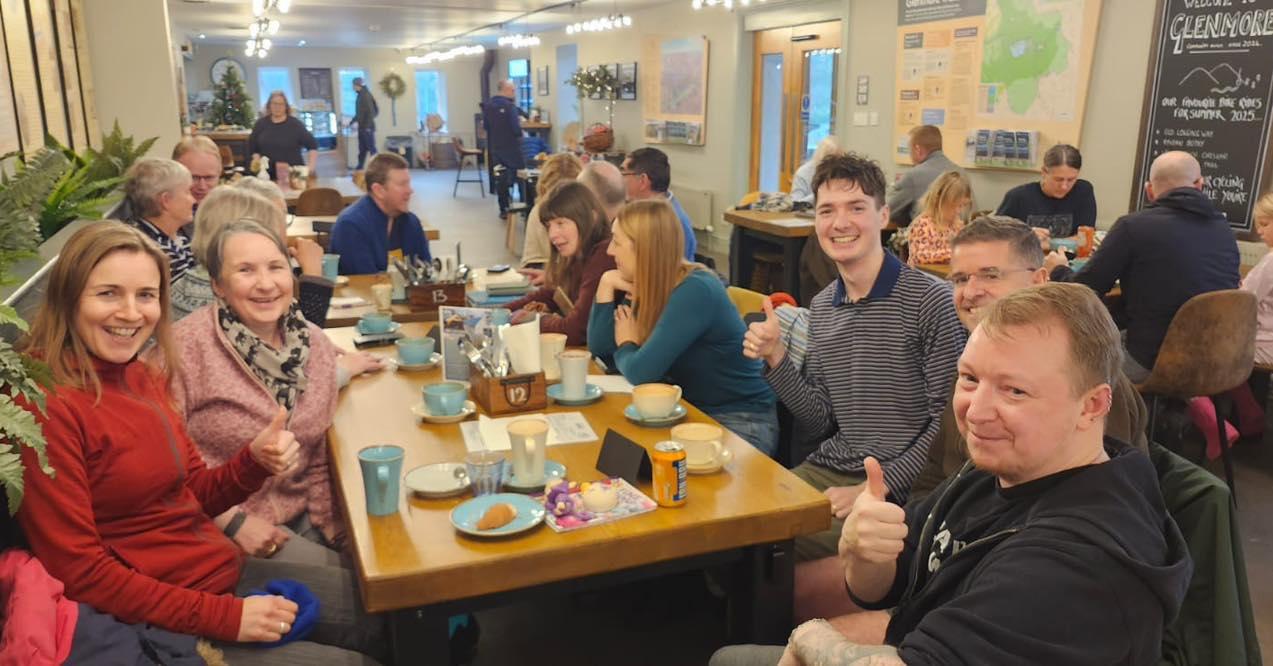 A group of people in a cafe smile at the camera, sitting around a table laden with coffee and food.