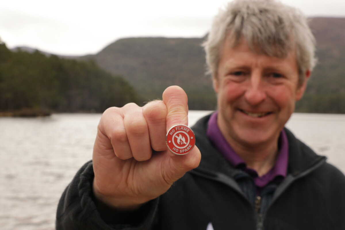 Person standing in front of a loch holding a badge in the foreground that says Protect our National Park. No flame, no spark. on it.