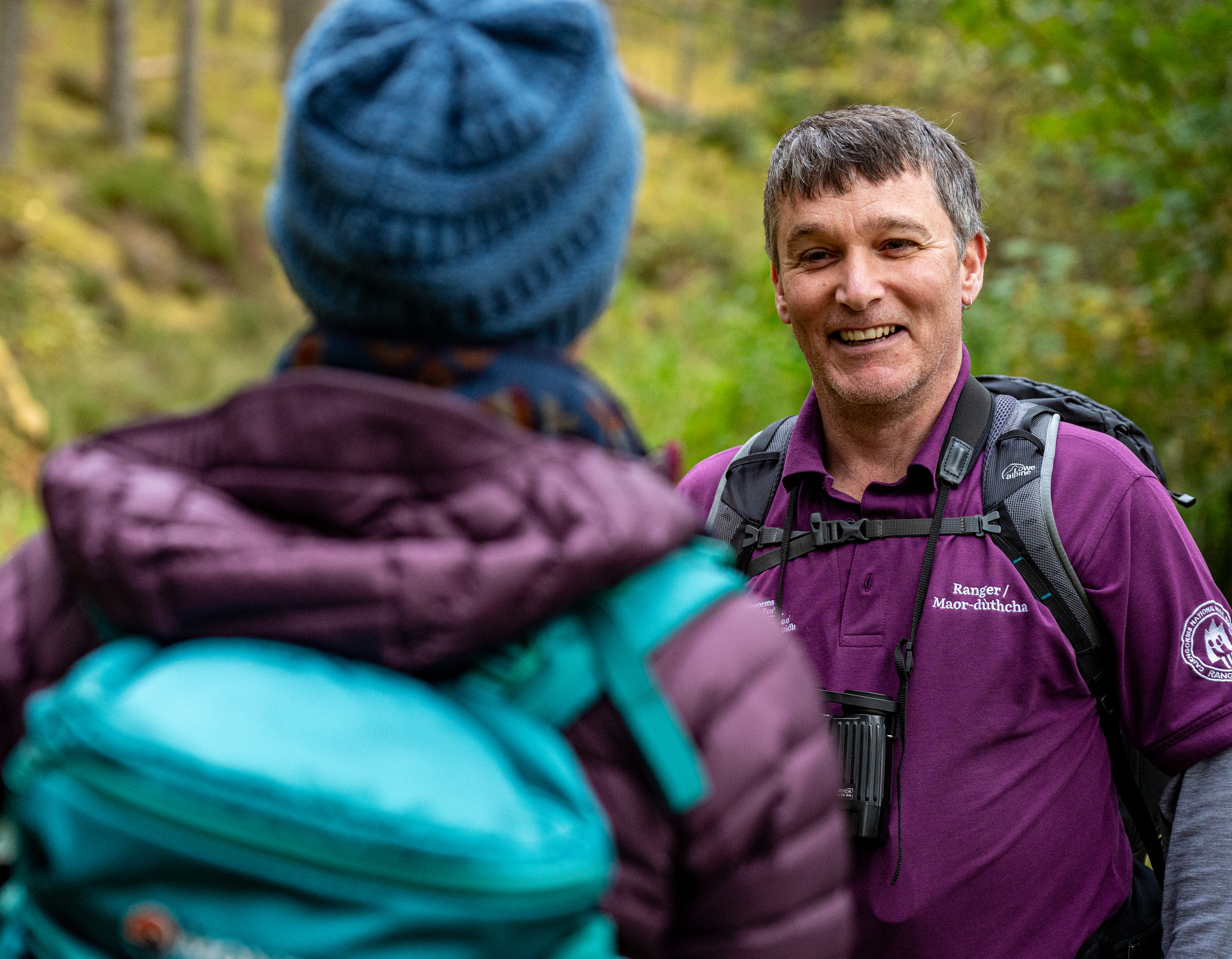 A Park Authority seasonal ranger wearing a purple polo shirt with binoculars and a black backpack talks to a visitor wearing a blue bobble hat, purple jacket and bright blue backpack.