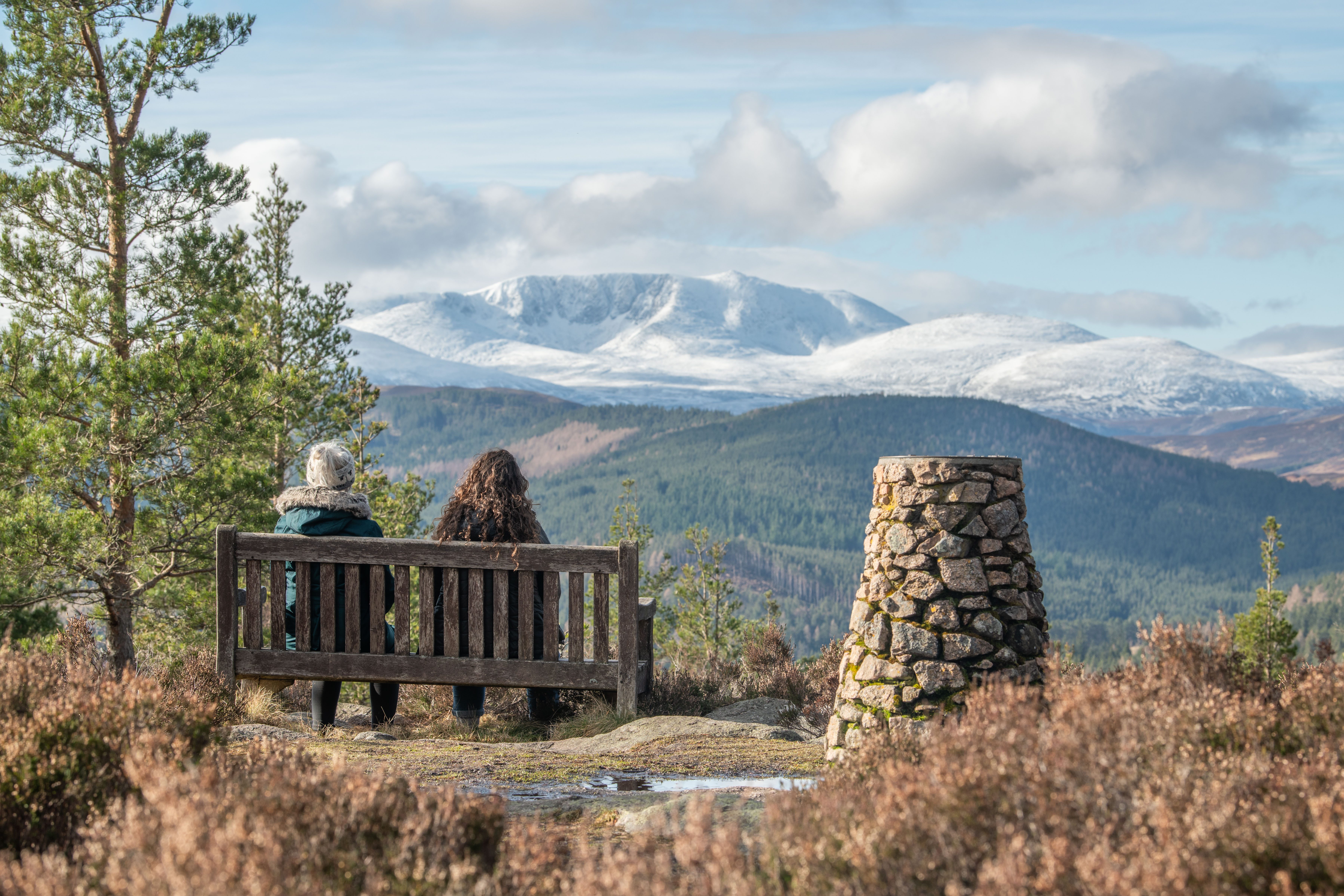 Two women sit on a wooden bench next to a stone cairn and some pine trees, looking out across snow-capped mountains