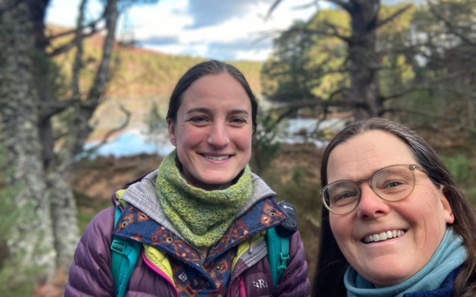 Two women smiling taking a selfie in the outdoors with trees and a loch in the background.