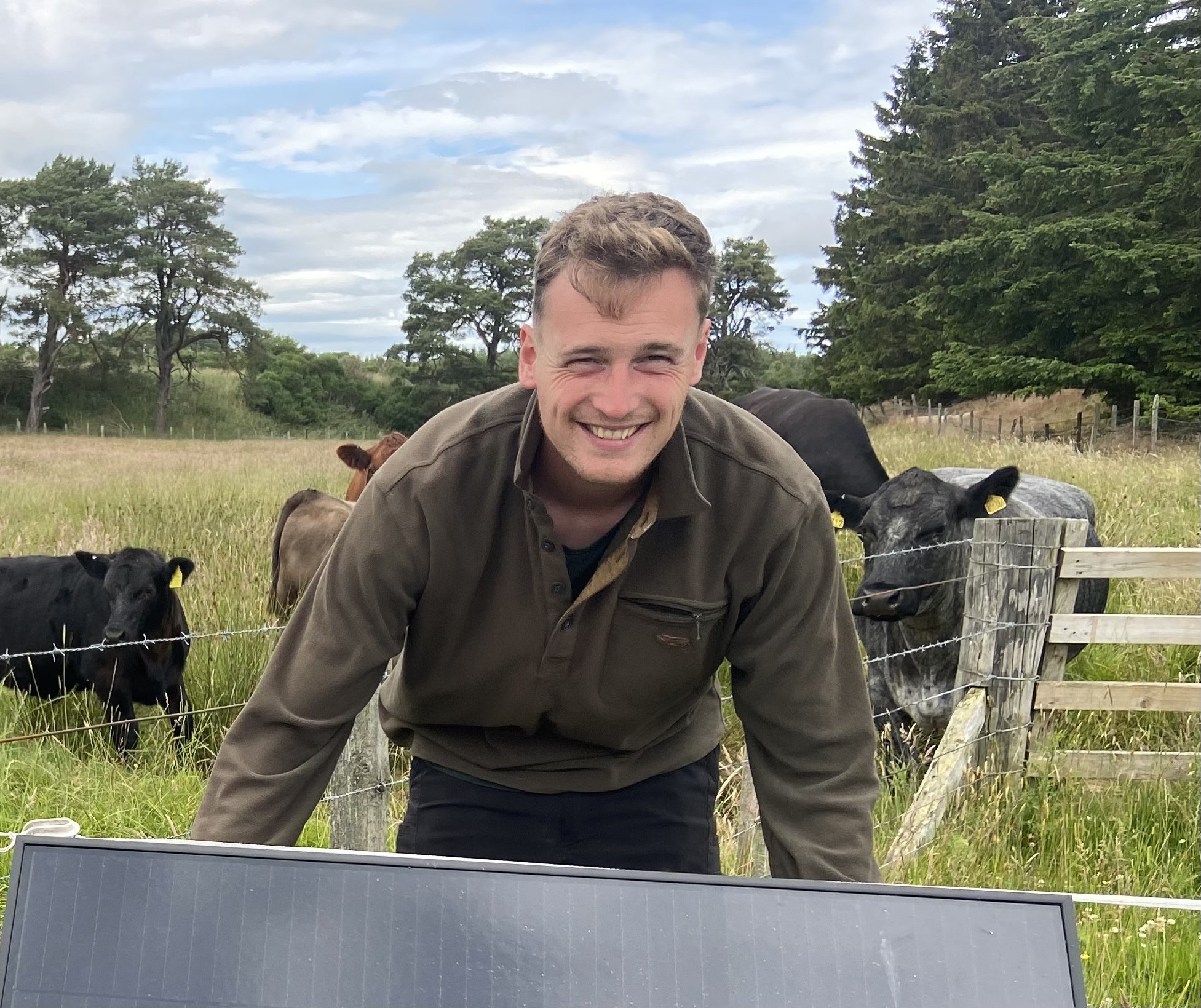 Young man smiles to camera whilst lifting heavy opject against a backdrop of lush fields and cows