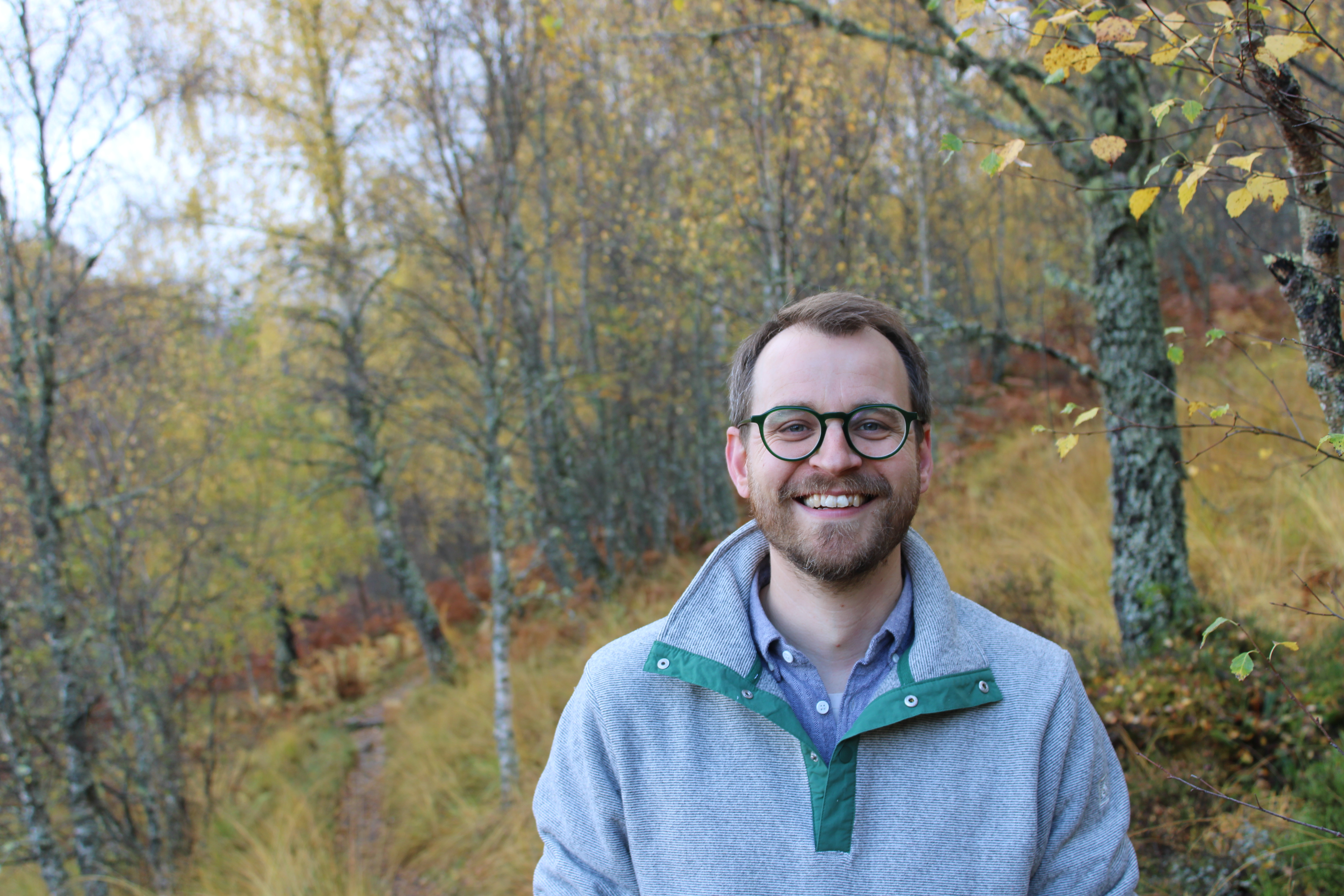 Olly stands smiling against a backdrop of autumnal leaves