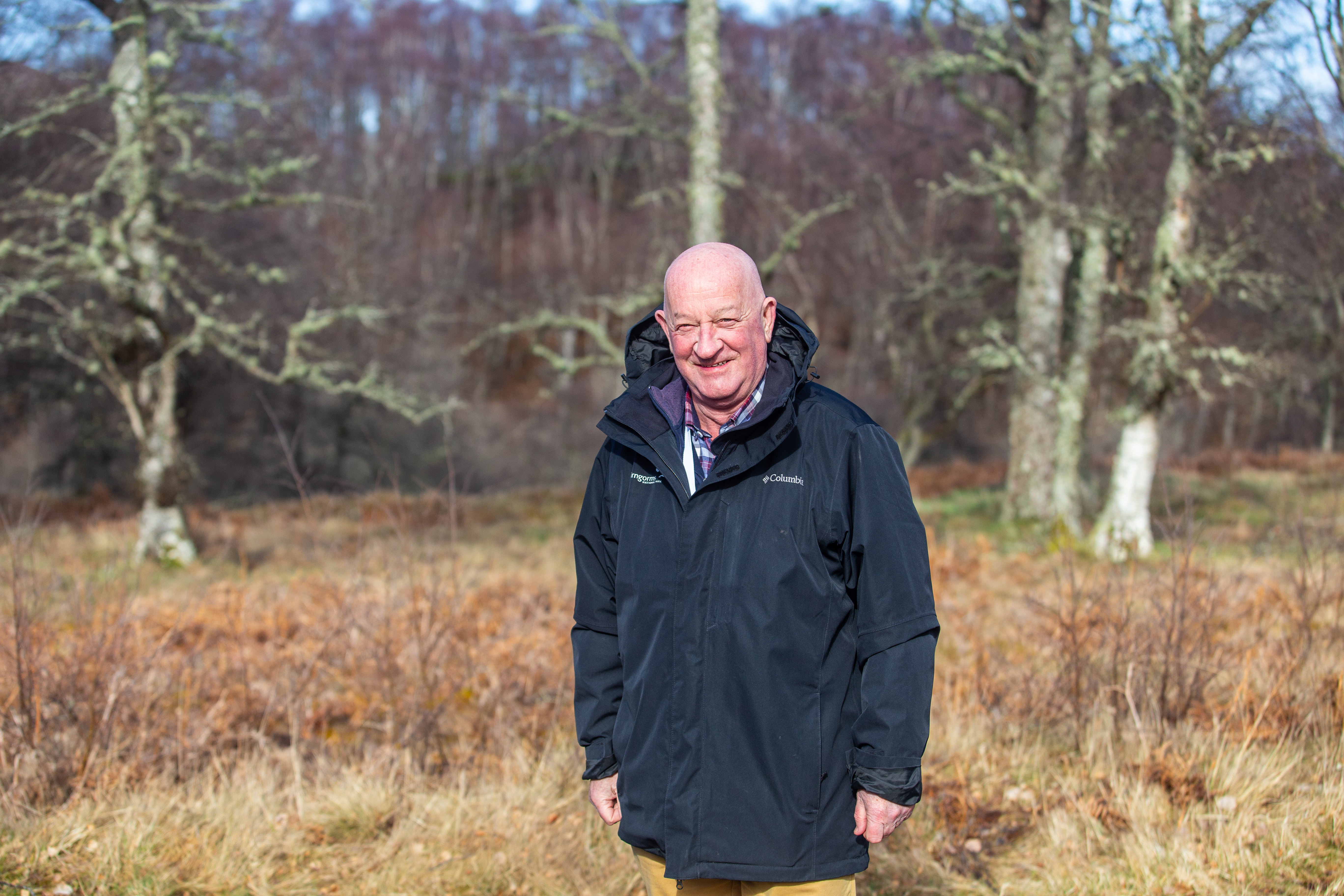 A man in a Park Authority jacket smiles at the camera in the sunshine against a backdrop of trees and bracken