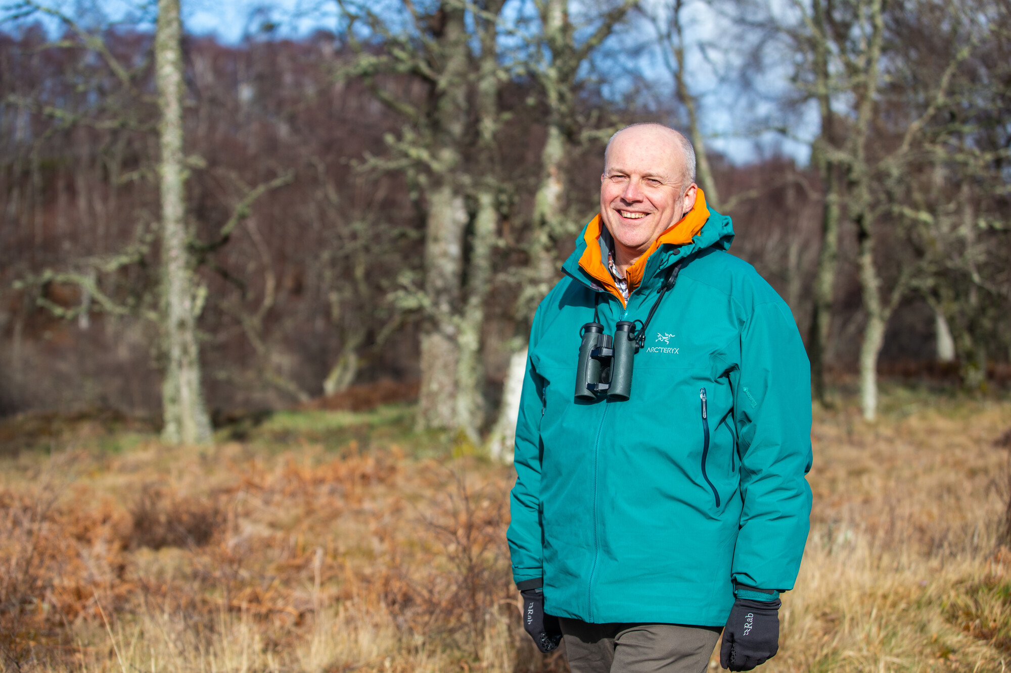 Man smiles to camera with binoculars around his neck, wearing a bright turquoise jacket