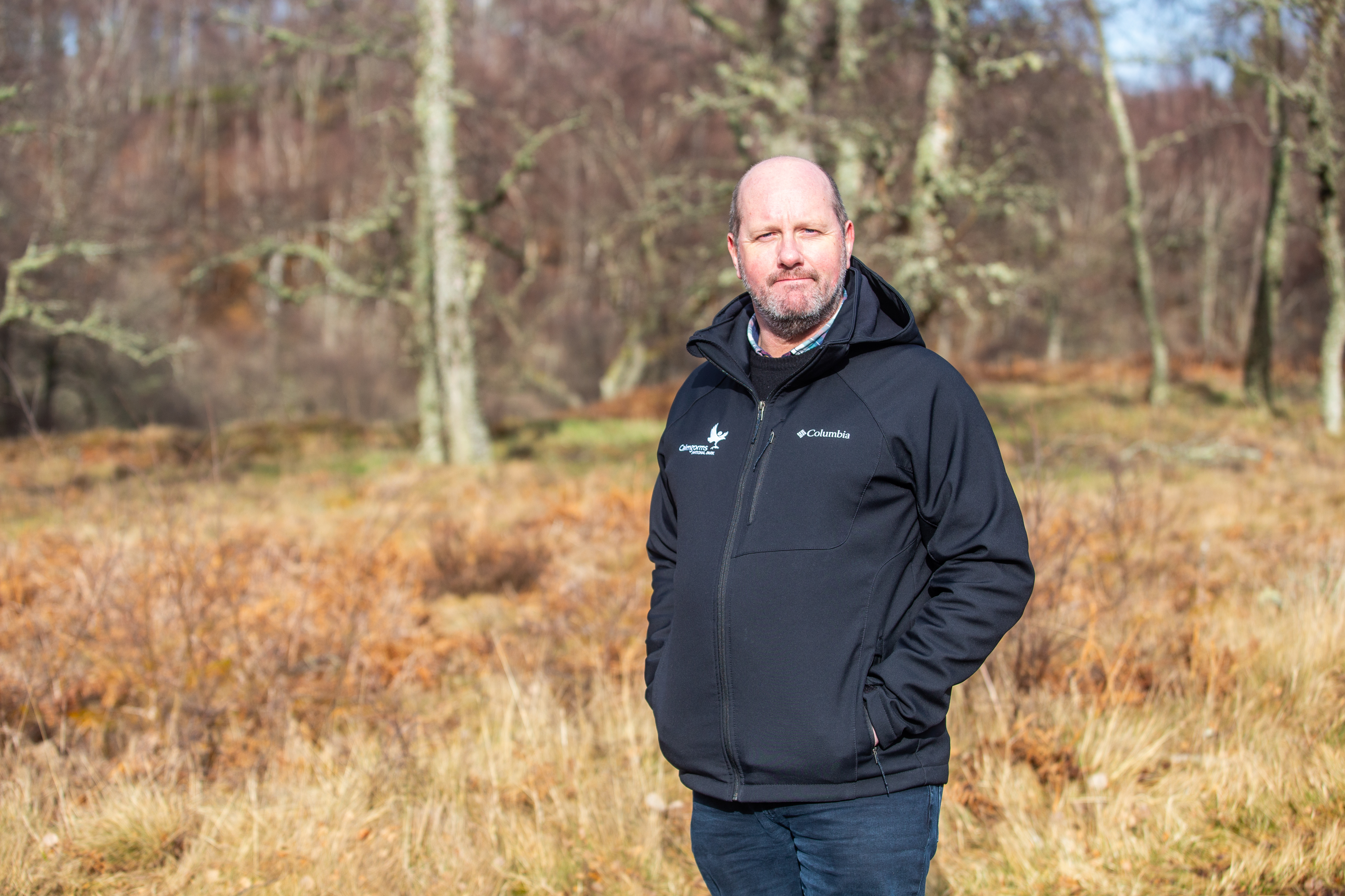 Chris stands against a backdrop of trees and bracken smiling to the camera in a National Park jacket