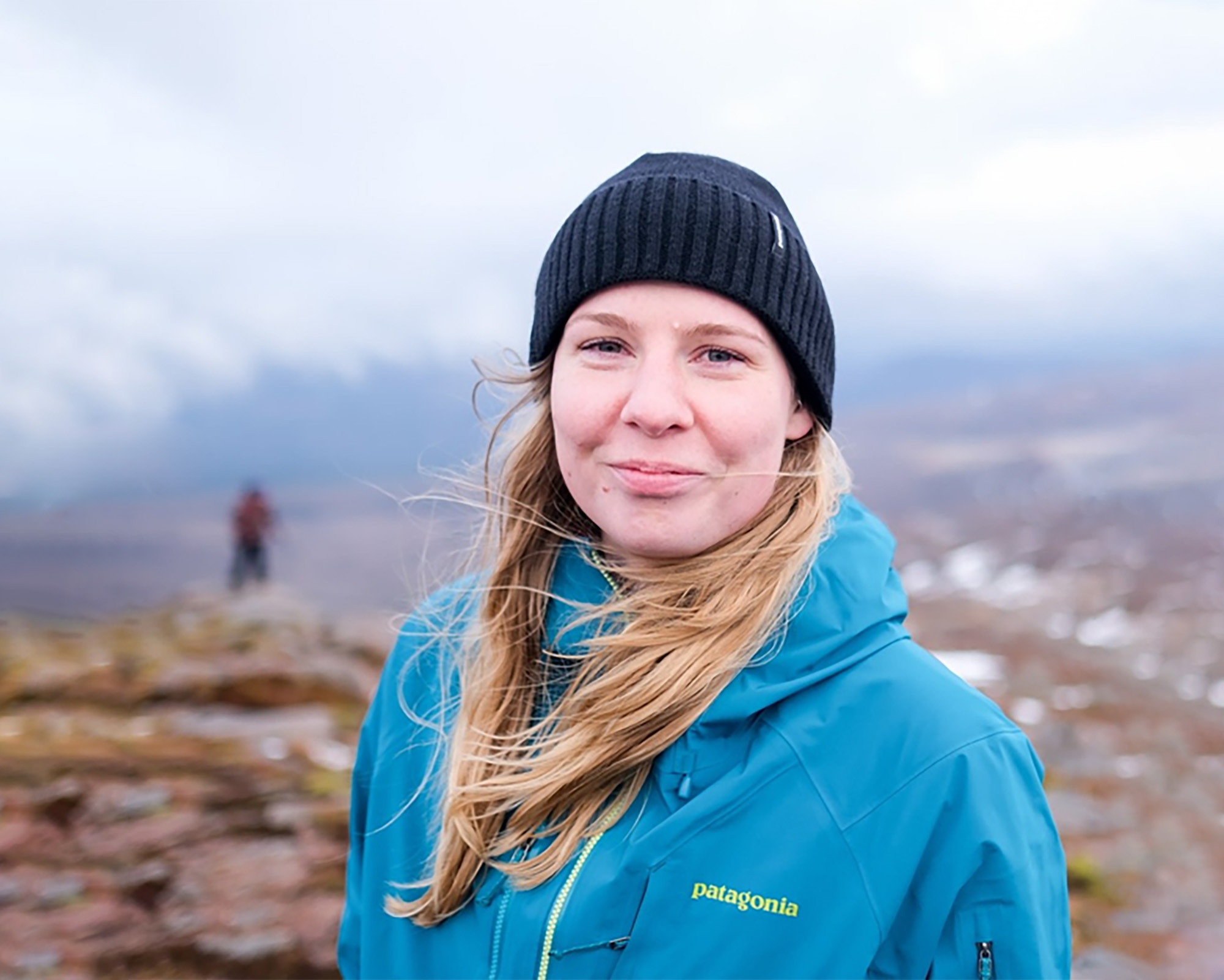 Woman smiles to camera in blue jacket with hat on against a backdrop of mountains