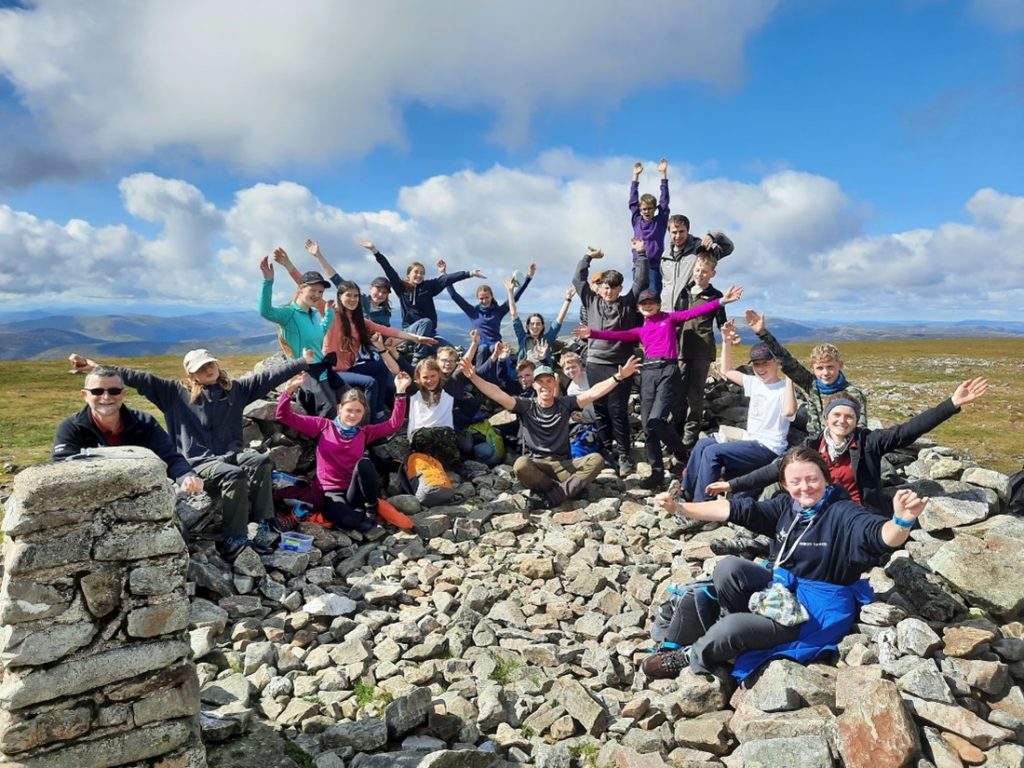 Junior rangers smile with their arms outstretched at the top of a hill with clouds and more hills in the background